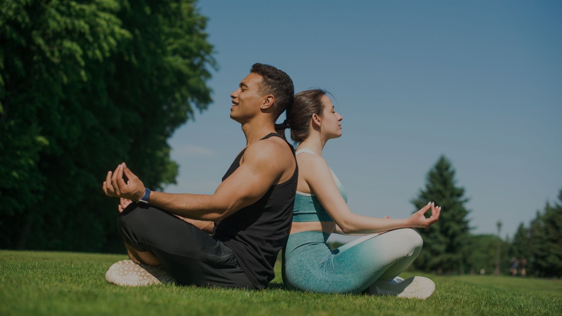 Two people meditating on grass with trees and blue sky in the background